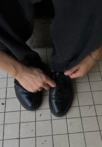 Mani che allacciano scarpe nere eleganti mentre indossa pantaloni scuri, seduto su un pavimento piastrellato con sfondo di pietra.