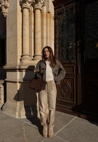 Žena v béžových nohaviciach, károvanom saku a čižmách drží šálku kávy a hnedú tašku, stojí pri vyrezávaných kamenných stĺpoch a drevených dverách.