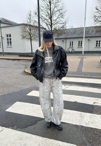 Veste en cuir noire portée sur un sweat-shirt gris avec un texte blanc, associée à un pantalon large gris clair à motifs et des bottes noires.