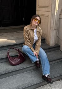 Rå støvjakke over en hvit topp, blå vide bukser, brune plattform loafers, gule solbriller og en burgunder vevd veske på trappen.