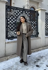 Γυναίκα με μακρύ καφέ παλτό και μπεζ πουλόβερ κρατάει ένα φλιτζάνι καφέ, στέκεται σε χιονισμένο έδαφος δίπλα σε περίτεχνο μαύρο μεταλλικό φράχτη.