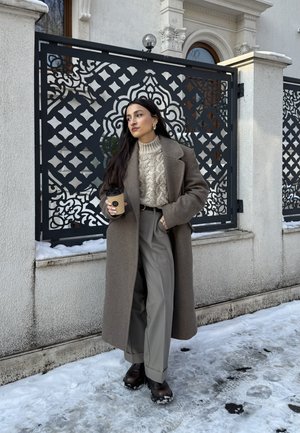 Mujer con un abrigo largo marrón y suéter beige, sosteniendo una taza de café, de pie sobre un suelo cubierto de nieve junto a una valla ornamental de metal negro.