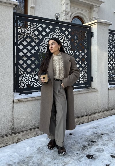 Mujer con un abrigo largo marrón y suéter beige, sosteniendo una taza de café, de pie sobre un suelo cubierto de nieve junto a una valla ornamental de metal negro.
