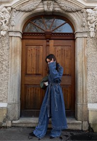 Dolga denim plašč z širokimi rokavi, zategnjenim pasom in gumbnim zapiranjem. Nosite ga preko ohlapnih denim hlač. Olivno zelena torba in skodelica kave v rokah.