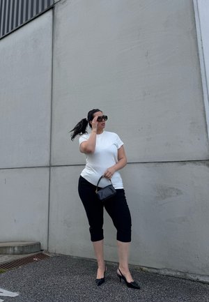 Mujer con gafas de sol, camiseta blanca, pantalones cortos negros y tacones, de pie junto a una pared de hormigón gris, sosteniendo un pequeño bolso negro.