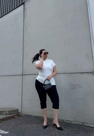 Mujer con gafas de sol, camiseta blanca, pantalones cortos negros y tacones, de pie junto a una pared de hormigón gris, sosteniendo un pequeño bolso negro.