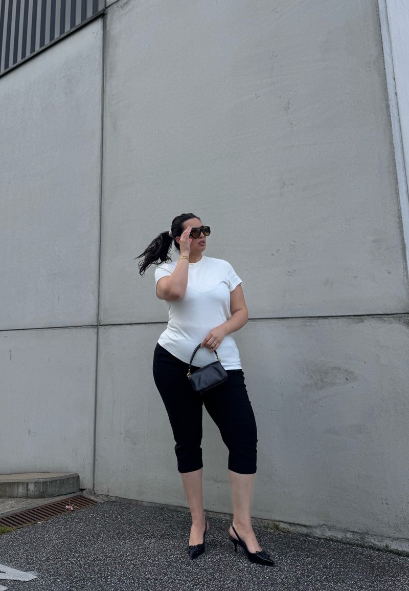 Woman wearing sunglasses, white t-shirt, black cropped pants, and heels stands beside a gray concrete wall, holding a small black handbag.