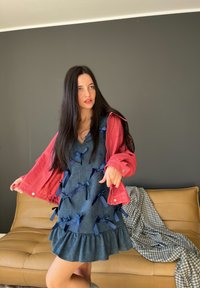 Vestido de denim con lazos navy, dobladillo acampanado y chaqueta rosa. Modelo posando en un sofá color beige, fondo gris y manta a cuadros cercana.
