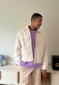 Lys beige glidelåsjakke med to frontlommer, brukt over en grovstrikket lilla genser, kombinert med lyse beige bukser. Jevn tekstur.