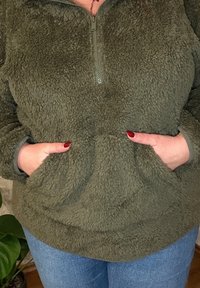 Sudadera de forro polar verde oliva con media cremallera, que cuenta con una tela suave y texturada y dos bolsillos frontales. Mangas largas con borde en puños.