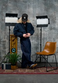 Personne portant une veste à col en fourrure, un large pantalon en denim et une casquette, debout sur un tapis à motifs entre des éclairages de studio, une plante et une chaise marron.
