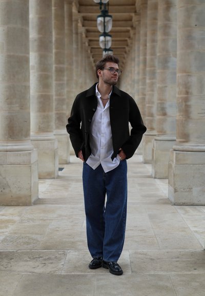 Chaqueta de tela negra con cuello, camisa blanca y vaqueros azul de corte holgado. De pie en un entorno arquitectónico con columnas de piedra y faroles.
