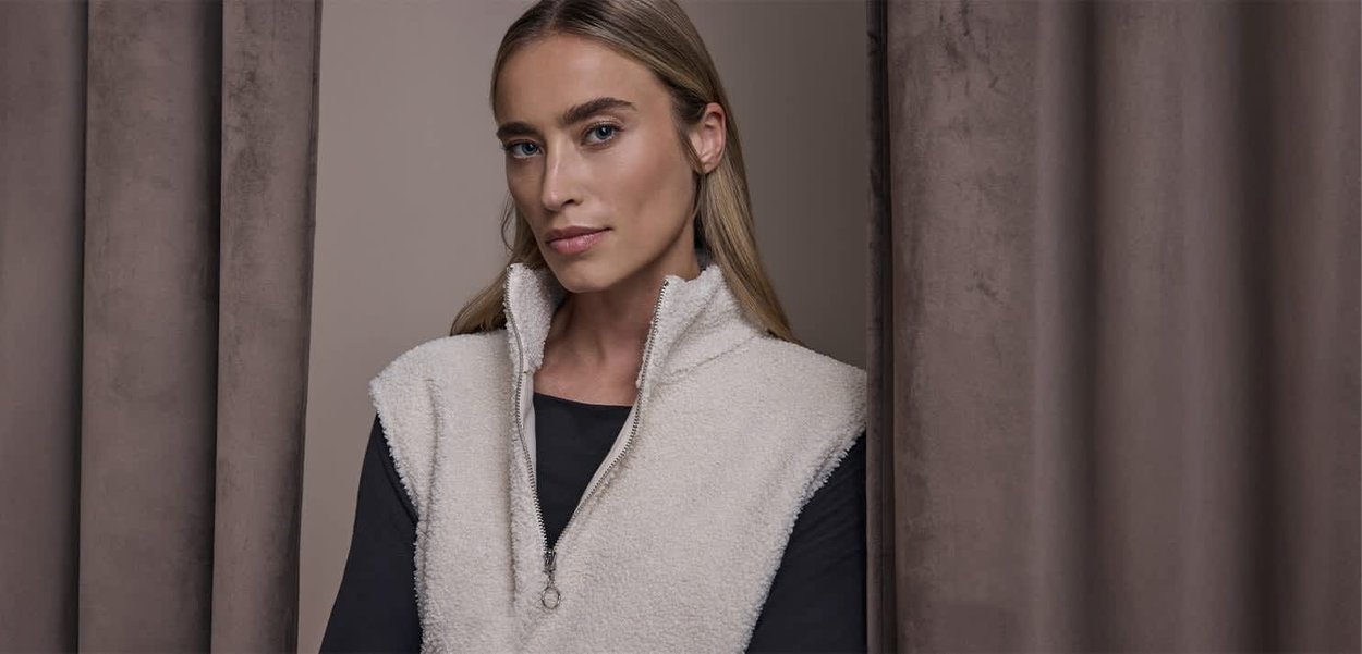 Gilet en polaire beige clair avec col montant et détail zippé, porté sur un haut noir, en arrière-plan neutre avec des drapés de tissu doux.