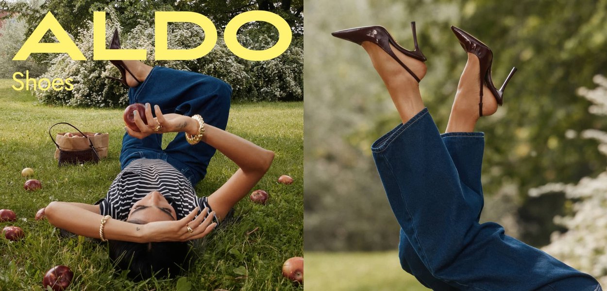 High-heeled shoes with a pointed toe and glossy dark brown finish, worn with wide, blue denim jeans on grass, surrounded by red apples.