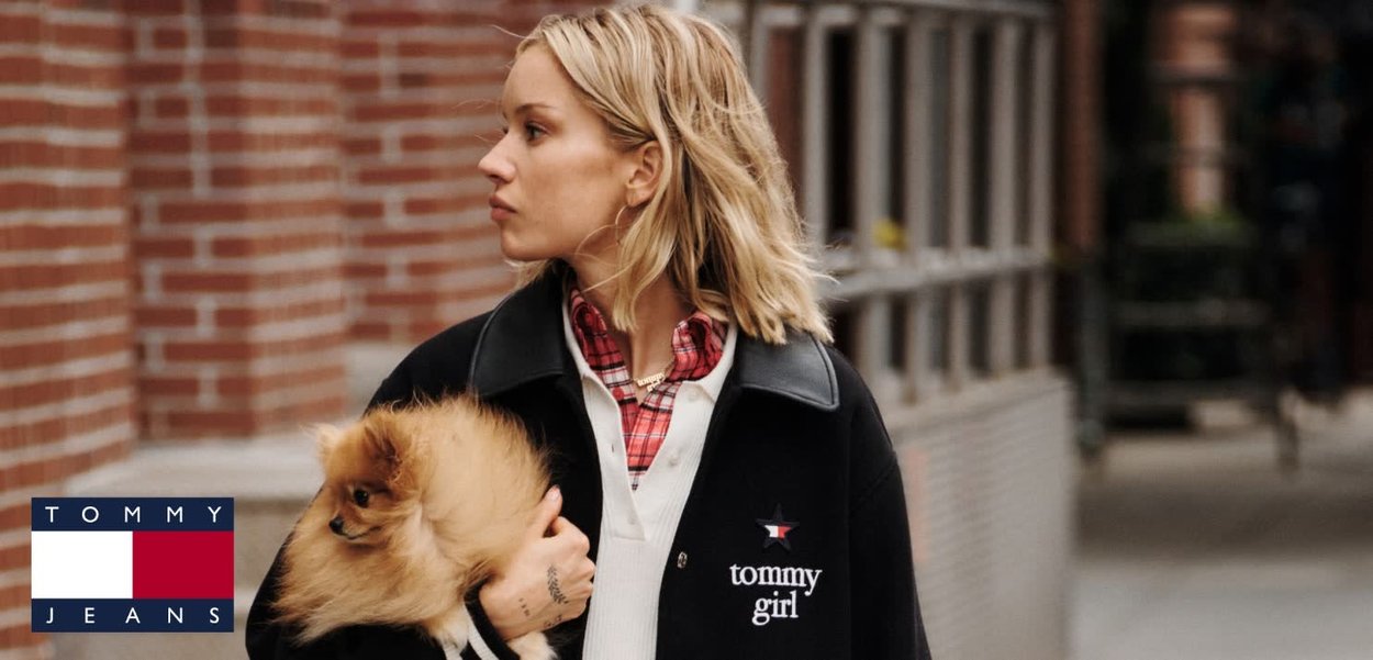 Abrigo negro con cuello, con bordado de "tommy girl". Superpuesto con una camisa de cuadros. Sosteniendo un pequeño perro tan esponjoso. Fondo de ladrillo.