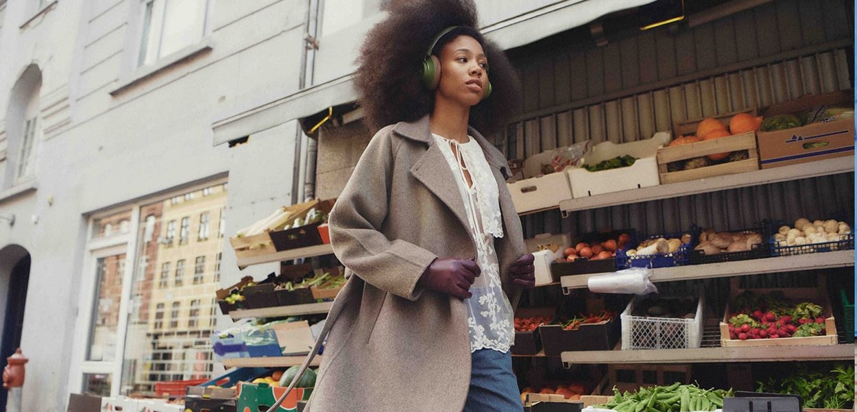 Gray wool coat over white lace top, blue jeans, purple gloves, and green headphones. Background features vegetable stands in a market.
