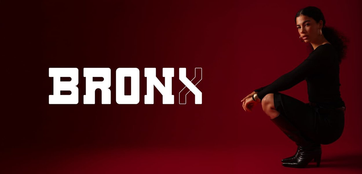 Schwarzes langärmliges Oberteil, knielanger Rock und kniehohe schwarze Stiefel. Modell mit dunklen Haaren posiert vor einem dunkelroten Hintergrund mit dem Schriftzug "BRONX".