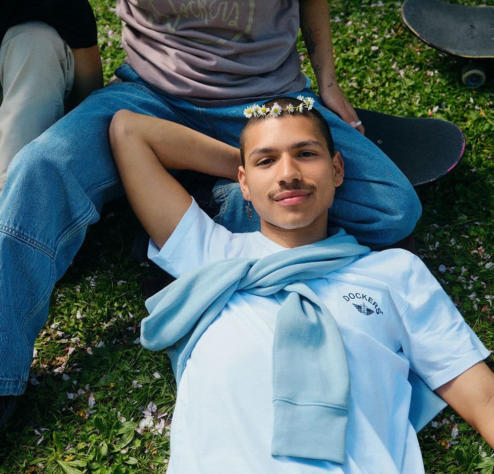 T-shirt bleu clair avec le logo "Dockers", pull bleu clair attaché autour du cou, couronne florale, assis sur l'herbe parmi des skateurs.