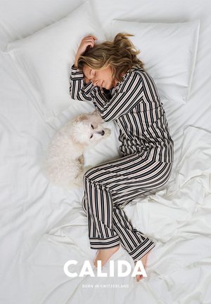 Gestreiftes Loungewear-Set in Schwarz und Creme, bestehend aus einem Langarmshirt und einer Hose. Ein fluffiger weißer Hund liegt neben einer Person auf weißen Bettwäsche.