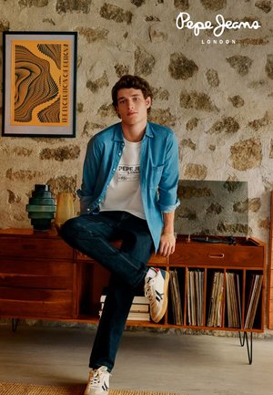 Spijkerjas over een wit T-shirt, donkere jeans en sneakers; staand naast een houten meubelstuk met kleurrijke vazen en platen.