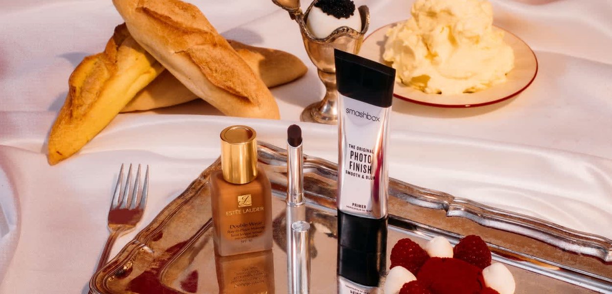Braune Glasflasche mit Estée Lauder Foundation, silberne Tube Smashbox Primer, Schokoladenriegel, Baguette, Sahne und Himbeer-Dessert auf einem Tablett.