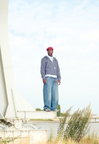 Een persoon staat op een metalen balk, gekleed in een grijze hoodie, blauwe wijde jeans en een rode pet. De achtergrond toont groen en water.