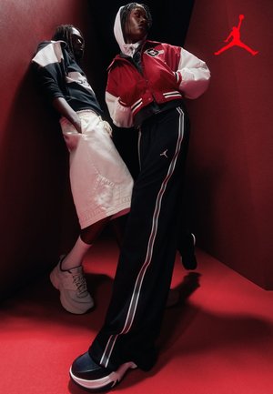 Dwie osoby ubrane w sportową odzież uliczną stoją w narożniku o czerwonym świetle, na ubraniach i butach widoczne są logotypy marki Jordan.