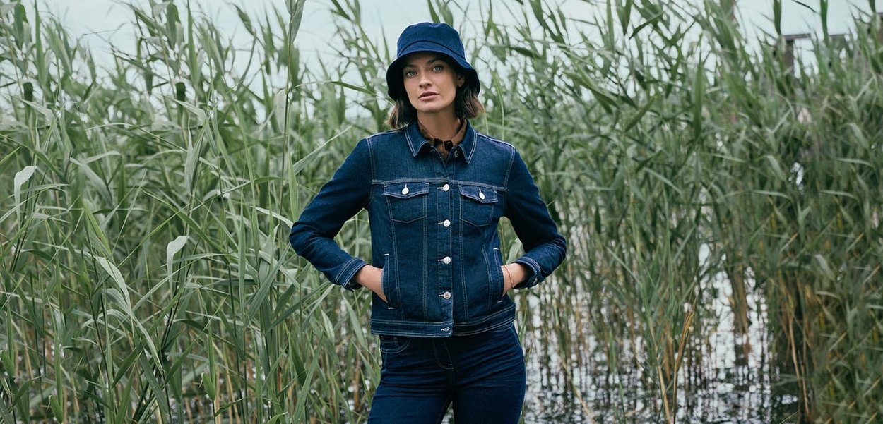 Jeansjacke und passende Jeans in Dunkelblau, mit silbernen Knöpfen und doppelten Brusttaschen, getragen mit einem blauen Bucket Hat, in einem mit Schilfrohr gefüllten Bereich.