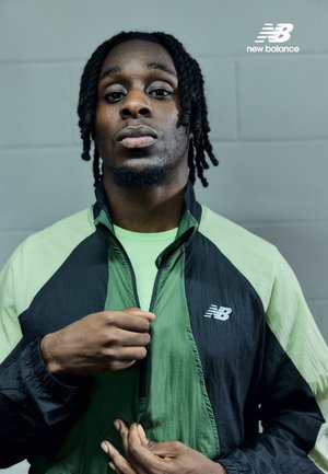 Chaqueta deportiva en tela negra y verde claro, con cremallera y un logo en el pecho. Material texturizado y diseño ajustado.