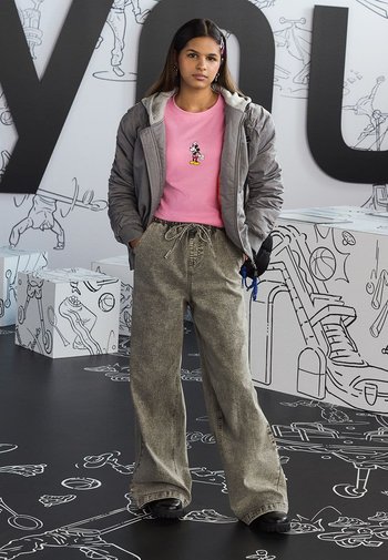Graue isolierte Jacke, pinkes T-Shirt mit Mickey Mouse, lockere graue Cordhosen, schwarze Schuhe, vor einem verspielten Hintergrund stehend.