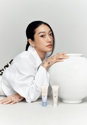 Zwei Hautpflegeröhrchen, eines beige und eines hellblau, neben einer großen weißen Keramikvase präsentiert. Modell mit langen schwarzen Haaren lehnt nachdenklich.