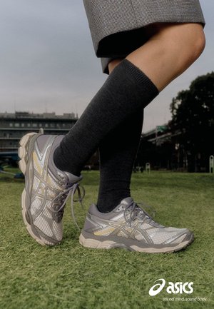 Ben klædt i grå knæstrømper og grå Asics løbesko står på græsset med en bylandskab i baggrunden.