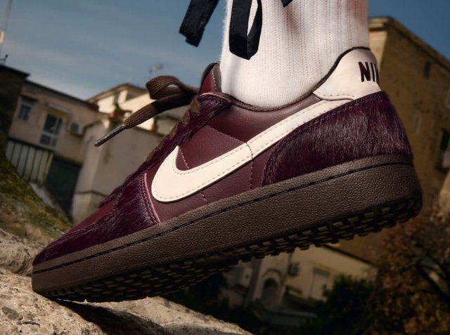 Bordeauxfarbene Sneaker mit fellartiger Textur, weißem Nike-Swoosh, strukturierter dunkler Sohle und schwarzen Schnürsenkeln; kombiniert mit einer weißen Socke.
