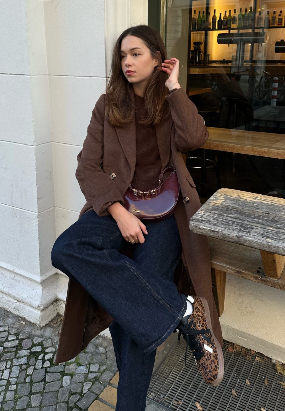 Abrigo de lana marrón sobre un suéter marrón, combinado con vaqueros de pierna ancha azul oscuro. Zapatillas con estampado de leopardo en marrón y negro y un bolso bandolera burdeos.