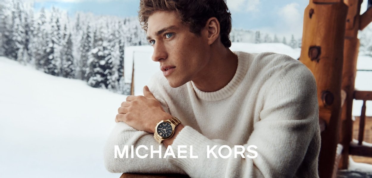 Gullfarget klokke med blå urskive, romertall og metallarmbånd, båret av en person i en hvit genser foran en snødekt bakgrunn.