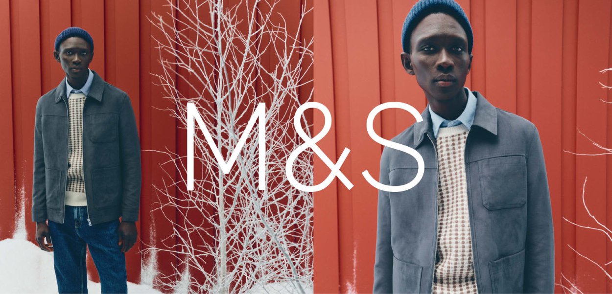 Szara zamszowa kurtka na swetrze w kratę z dzianiny i niebieska czapka z dzianiny, na tle czerwonej ściany z białymi gałązkami. Wyraźne logo M&S.
