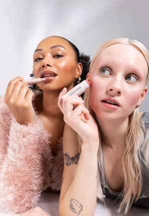 To kvinner holder skjønnhetsprodukter: en med en rosa, fluffy, oversized genser og en leppeglans, den andre med en sølvfarget topp og en hvit multistift.