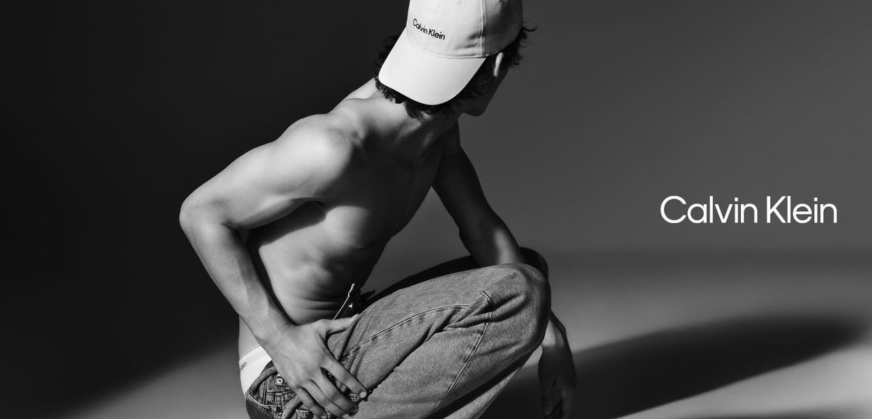 Una figura a petto nudo è accovacciata, indossando jeans di denim chiaro e un cappellino bianco con "Calvin Klein" ricamato in nero. L'immagine è in bianco e nero.