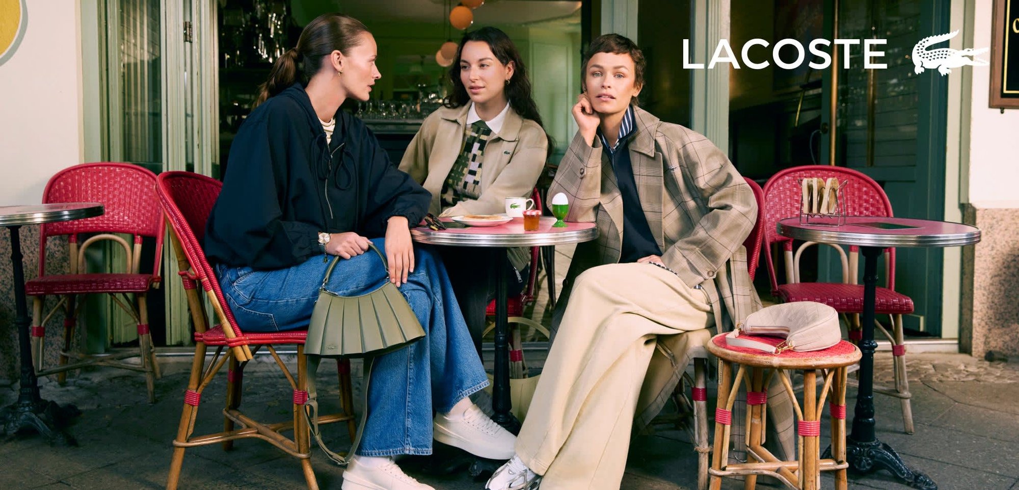 Trois femmes assises à une table dans un café. L'une porte une veste bleu marine et un jean, l'autre un manteau à carreaux beige, et la troisième des pantalons clairs et des baskets.