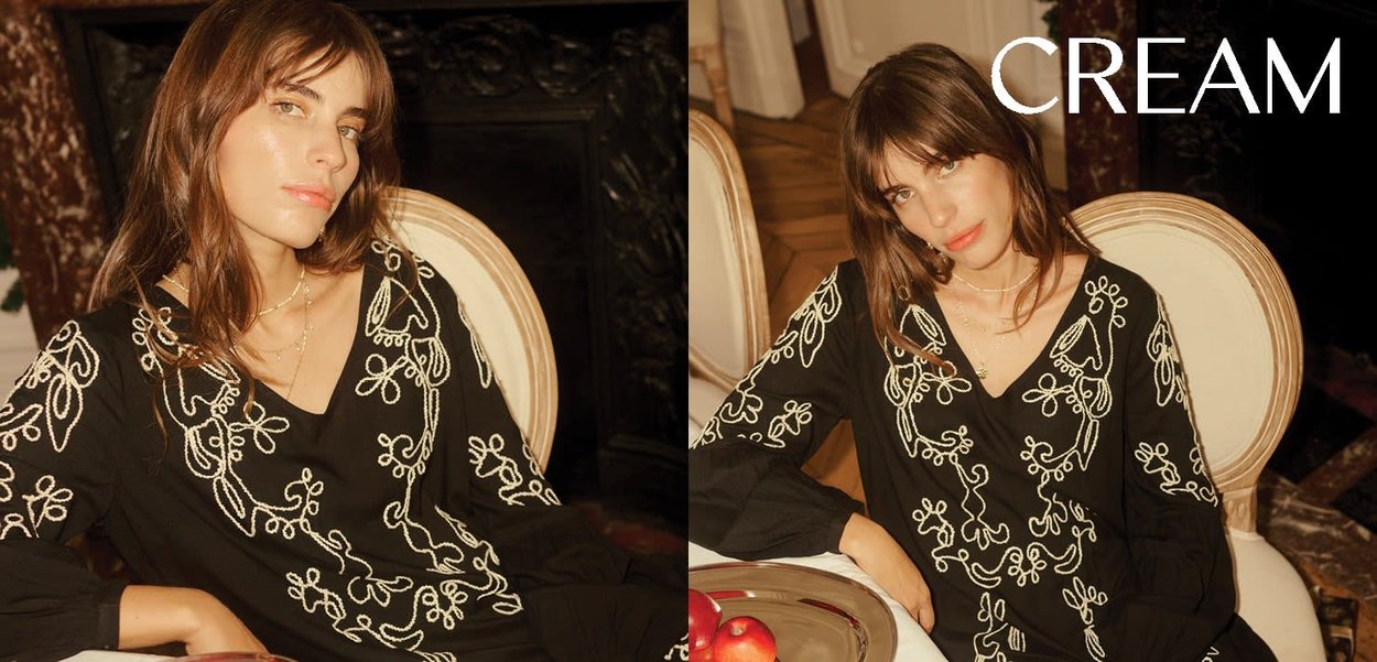 Black blouse with white floral embroidery, v-neckline, and long sleeves. Features soft texture and relaxed fit. Set in a well-decorated room.