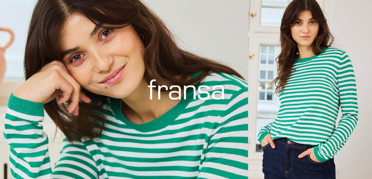 Junge Frau mit langen braunen Haaren, die ein grün-weiß gestreiftes Langarmshirt und dunkle Jeans trägt, posiert drinnen am Fenster.