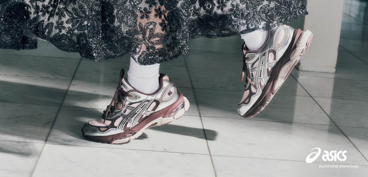 Persona con falda de encaje negra con estampado, calcetines blancos y zapatillas Asics rosas saltando sobre un suelo blanco con baldosas.