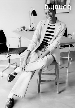 Blazer gris clair sur un haut rayé noir et blanc, associé à un jean en denim clair et des chaussures sombres élégantes. Assis sur un tabouret en bois.