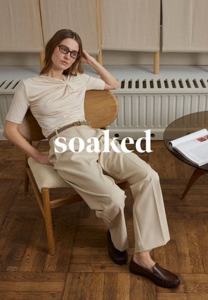 Frau trägt beigefarbenes Oberteil und Hose, sitzt entspannt auf einem Holzstuhl neben einem runden Tisch mit offener Zeitschrift auf dem Parkettboden.