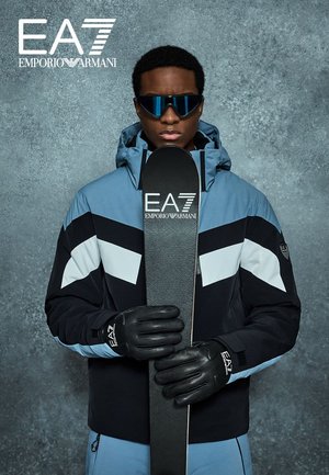 Blaue und schwarze Skijacke mit weißen Akzenten, Skihandschuhe und schwarzes Snowboard mit EA7-Logo. Elegante Sonnenbrille vervollständigt den Look.