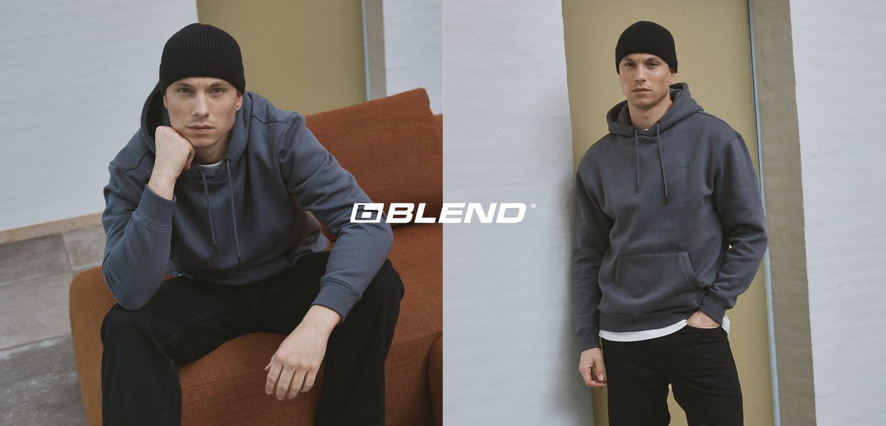 Sweat à capuche gris avec cordons, porté par une personne assise sur un canapé orange, associé à un jean denim foncé. Un bonnet noir complète le look.