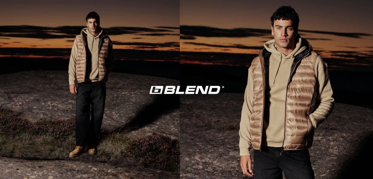 Beigefarbene gepolsterte Weste mit glänzender Oberfläche, getragen über einem beigen Hoodie und schwarzen Hosen. Das Modell steht auf felsigem Gelände vor einer Sonnenuntergangskulisse.