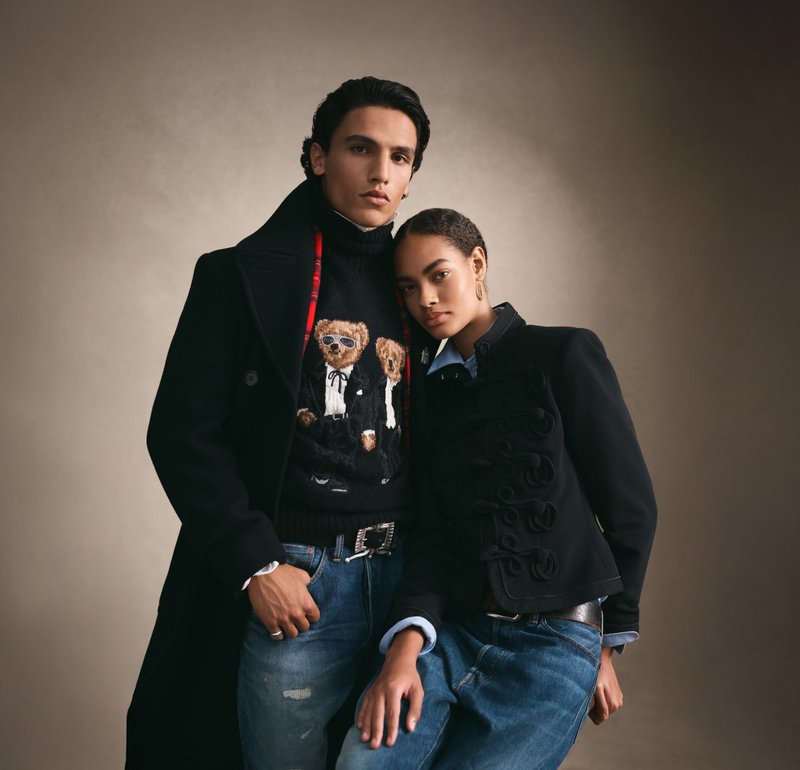 Joven y mujer con abrigos negros a la moda y vaqueros posando juntos contra un fondo neutro, el hombre lleva un suéter con diseño de oso de peluche.