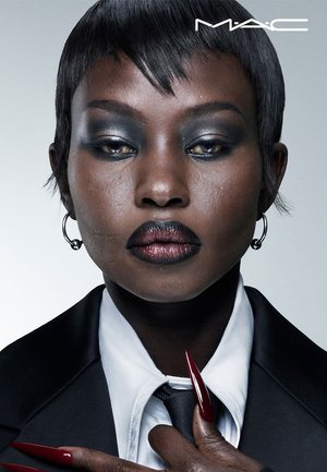 Model mit kurzen schwarzen Haaren, trägt einen maßgeschneiderten schwarzen Anzug und Krawatte. Dunkles Augen-Make-up, kräftige rote Lippen und lange rote Nägel. Silberne Creolen.