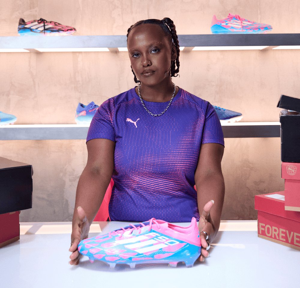 Puma Fußballschuhe in Pink und Blau, mit strukturiertem Design und schlanker Form, präsentiert auf einem Tisch mit Kisten im Hintergrund.
